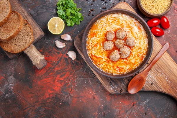 overhead view of meatballs soup with noodles uncooked pasta cutting board lemon a bunch of greens tomatoes different spices on dark table - Советы по выбору рецептов