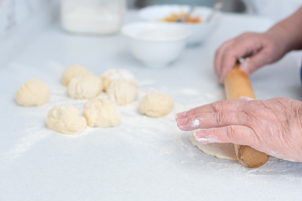 senior woman hands rolling out the dough with a rolling pin on a white kitchen table with blurred grated apple and sugar on background selective focus process of making pies with apple filling - Шанежки на кефире с картошкой