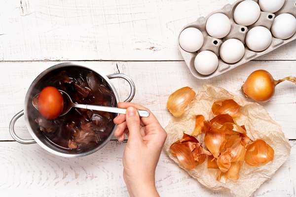 easter eggs the process of coloring with natural dye onion husks in a small saucepan on a white wooden table and a woman s hand holding a colored egg in a spoon - Яйца с рисунком и узорами в луковой шелухе