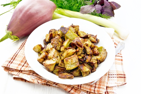 fried eggplant with onion egg and spices in a plate on a napkin basil leek and dill on white wooden board - Баклажаны, тушённые со свежими помидорами и луком