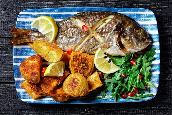 roasted dorado fish with baked potato coated in breadcrumbs served on a plate with a lemon wedge and fresh wild rocket salad with pomegranate seeds on a wooden background top view close up - Печёная рыба, постный стол