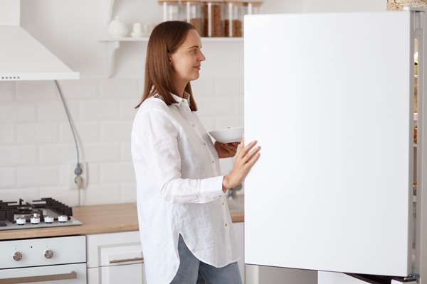 profile portrait of beautiful young adult woman wearing white shirt looking smiling inside fridge with pleasant smile holding plate in hands posing with kitchen set on background - Замороженные подосиновики