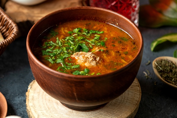 side view of traditional russian or ukrainian red soup borscht with beef and vegetables beet potato carrot cabbage onion fresh herbs and spices in clay bowl on bla - Харчо