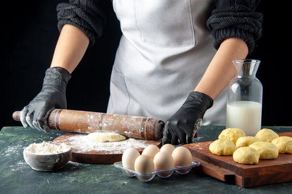 front view female cook rolling out dough with flour on dark cake job oven hotcake dough pie worker egg cuisine - Пирожки дрожжевые печёные