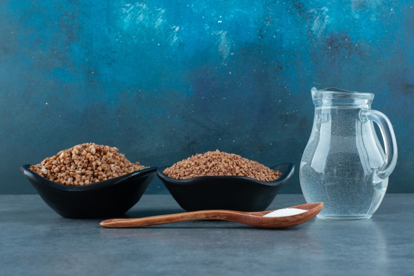 cooking ingredients of two bowls of buckwheat a small jug of water and a spoonful of salt on blue background high quality photo - Гречневое молоко