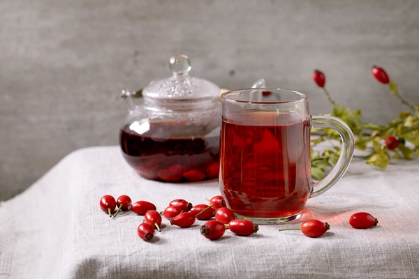 transparent cup of rose hip berries herbal tea and glass teapot standing on white linen table cloth with wild autumn berries around winter hot cozy beverage - Напиток из шиповника (школьное питание)